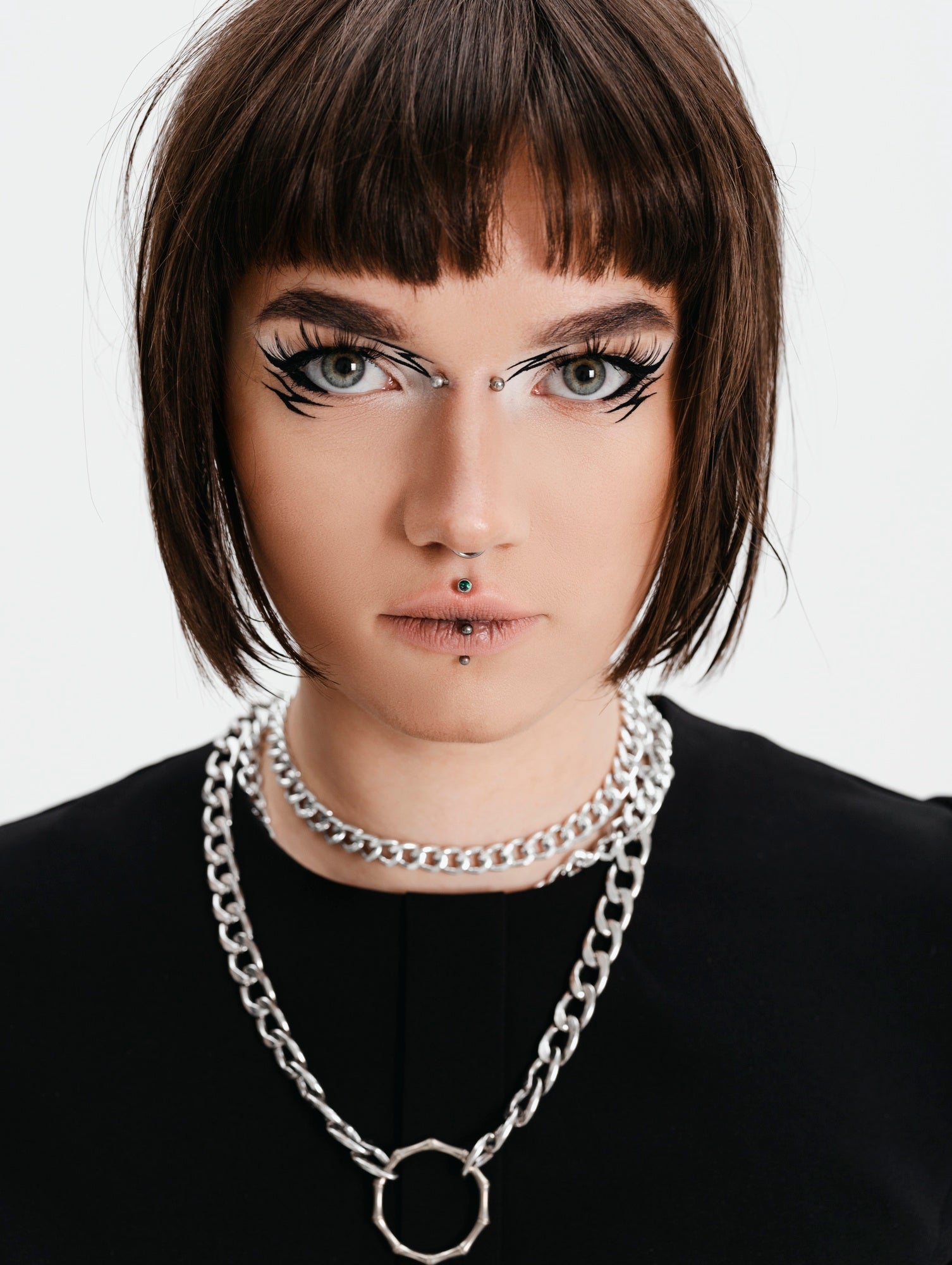 Person with dramatic makeup and a bob haircut wearing a black top and silver chain necklace.