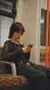A person with short dark hair wearing a black mesh top and layered accessories sits on a patterned train seat, looking down at their phone while riding public transport.