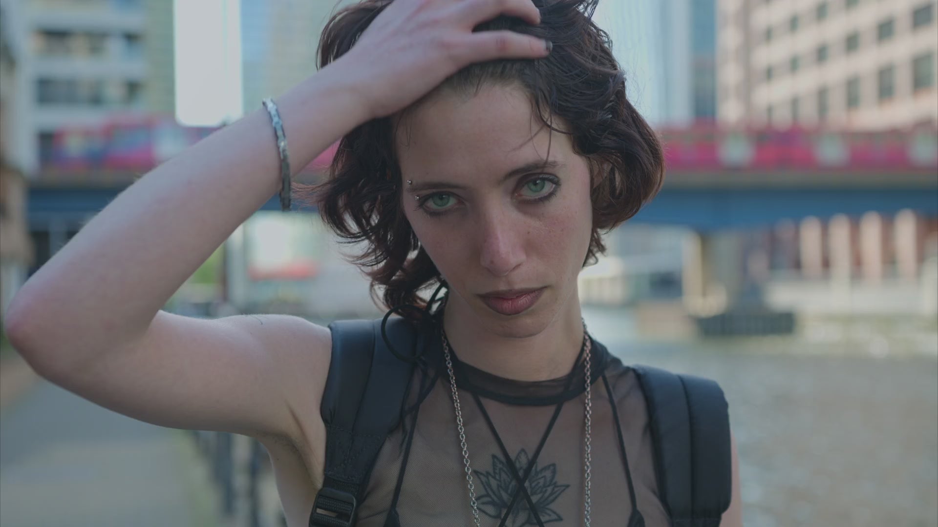 A person with short dark hair stands outdoors in an urban setting, lifting a hand through their hair and looking directly at the camera. They are wearing a sheer black top, layered necklaces, and a backpack, with tall buildings and a bridge blurred in the background.