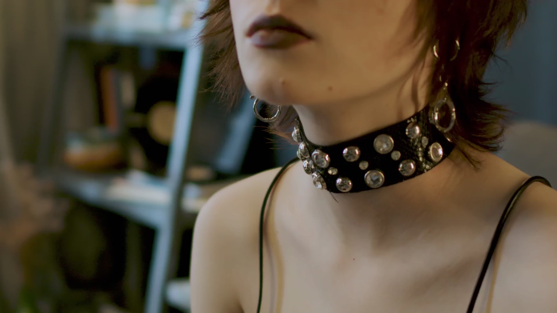 A close-up of a person wearing a black leather choker decorated with large silver studs and rhinestones, paired with dark lipstick and silver earrings, shown from the neck and shoulders upward in an indoor setting.